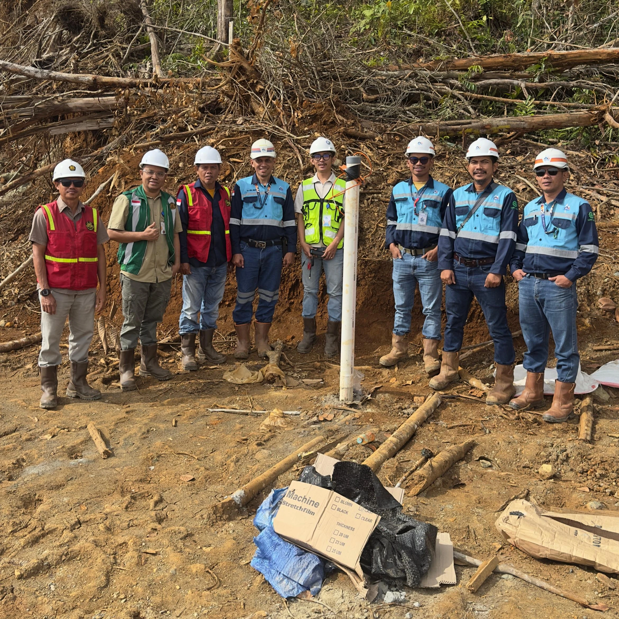 Foto grup tim instalasi dan pemantauan Piezometer dengan APD lengkap di lokasi tambang PT Mega Haltim Mineral, Kalimantan, berdiri di samping casing piezometer di area lereng hutan.