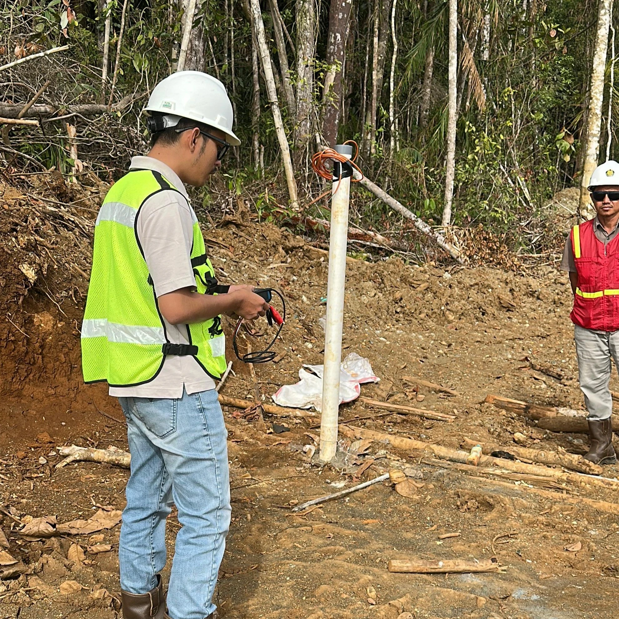 Seorang teknisi di lokasi tambang Kalimantan menggunakan pembaca manual (readout) untuk memantau data piezometer, dengan kabel terhubung ke pipa instalasi di lapangan terbuka.