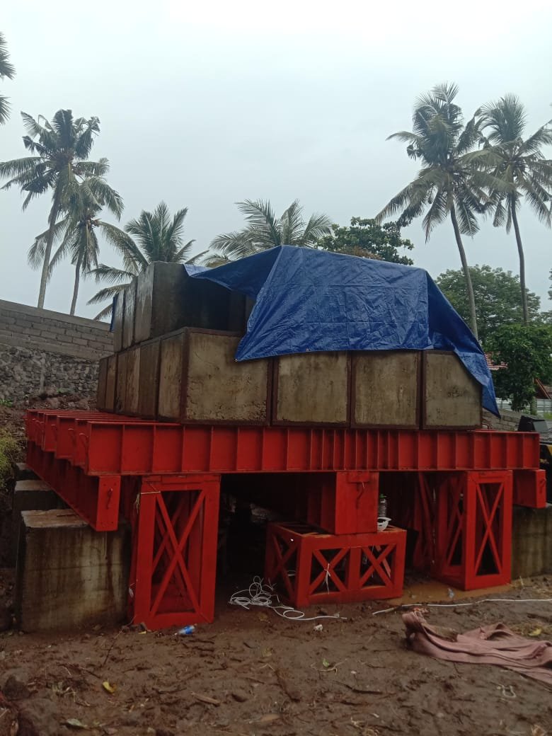 Struktur rangka baja oranye menahan tumpukan balok beton masif yang sebagian tertutup terpal biru.