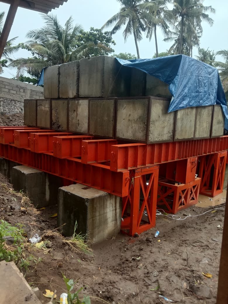 Tumpukan balok beton uji beban Kentledge di atas platform baja oranye dengan latar belakang pohon kelapa.
